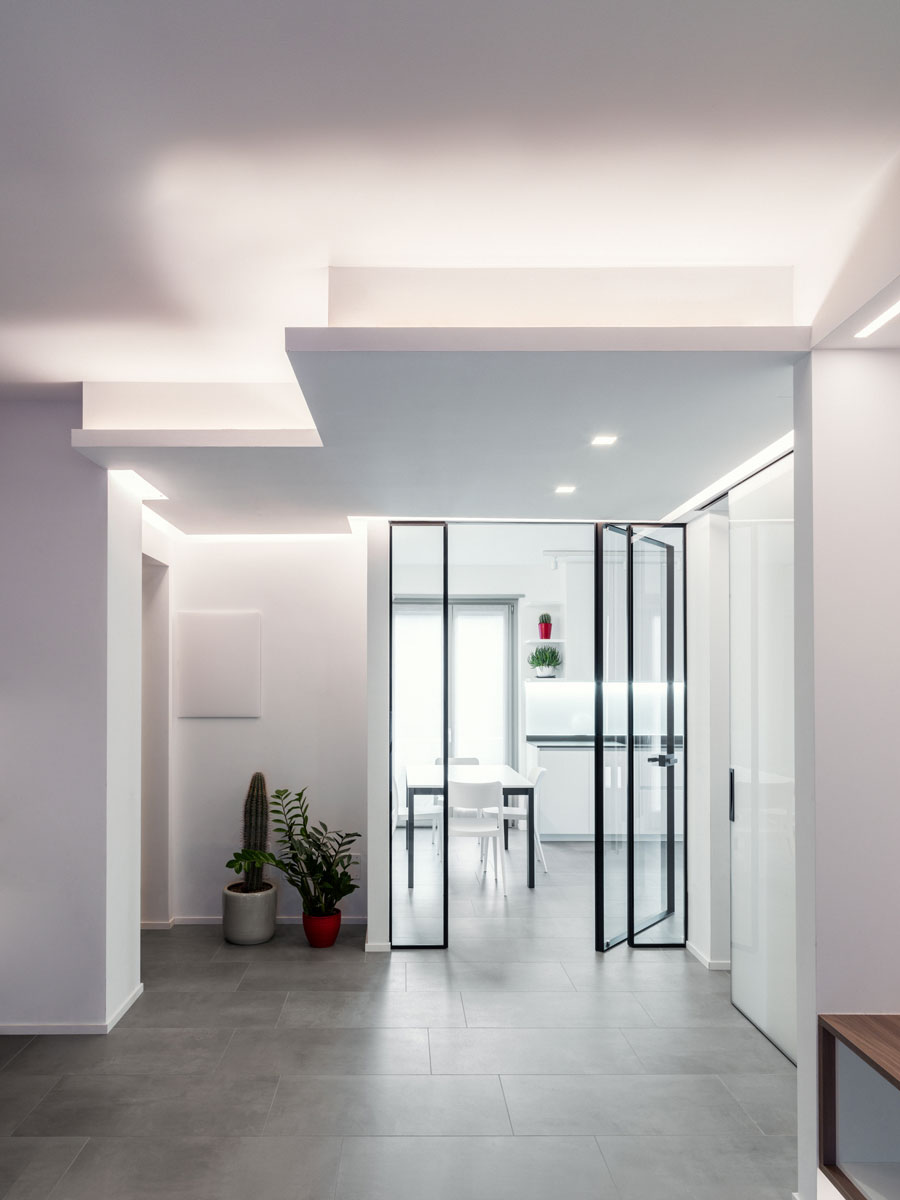 Stanza con illuminazione a soffitto e dietro una porta in vetro una cucina sullo sfondo