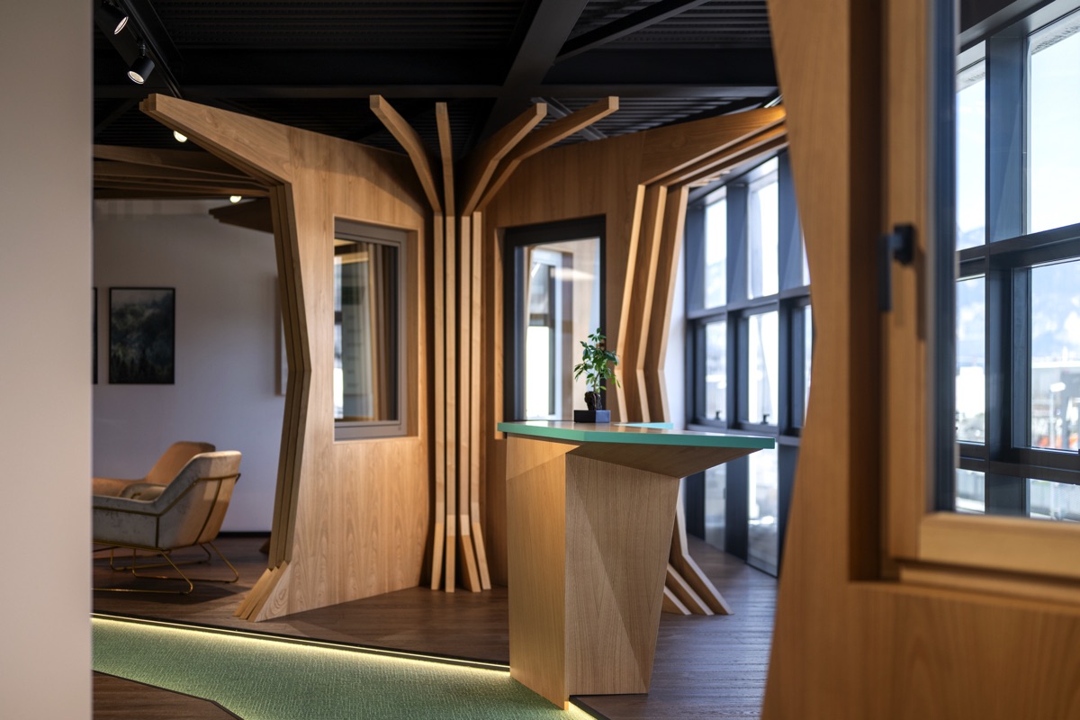 Un interno moderno con strutture in legno che ricordano tronchi di alberi. La stanza ha grandi finestre che permettono molta luce naturale. Un tavolo di design unico con una pianta sopra è presente al centro. Sullo sfondo si intravede una poltrona e un quadro.