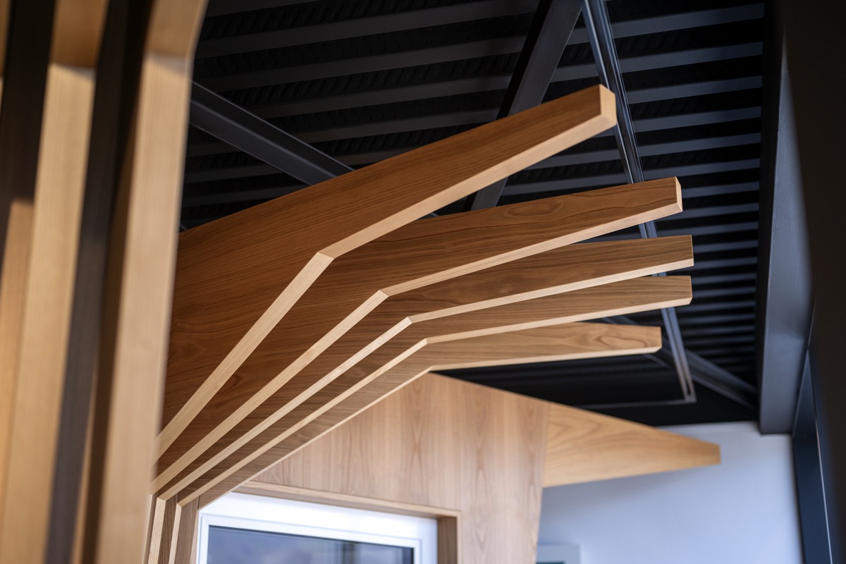 Dettaglio di una struttura in legno che ricorda i rami di un albero, con travi che si estendono dal centro verso l'esterno. La struttura è posizionata sotto un soffitto nero con travi in acciaio.