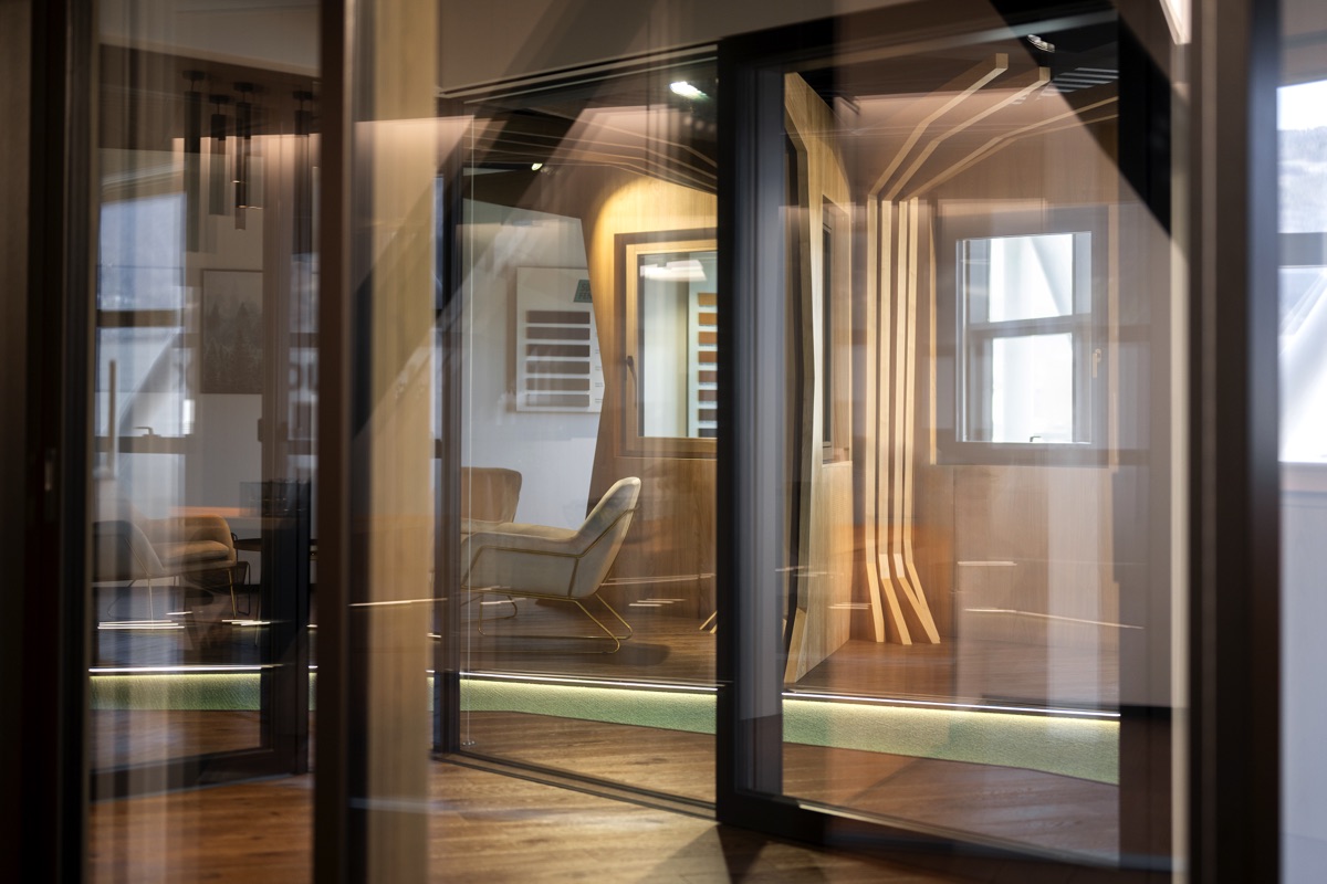 Vista di un interno moderno con pareti di vetro, mostrando vari spazi interni e riflessi. Un’area lounge con poltrone beige è visibile attraverso le pareti di vetro, evidenziando il design aperto e trasparente.