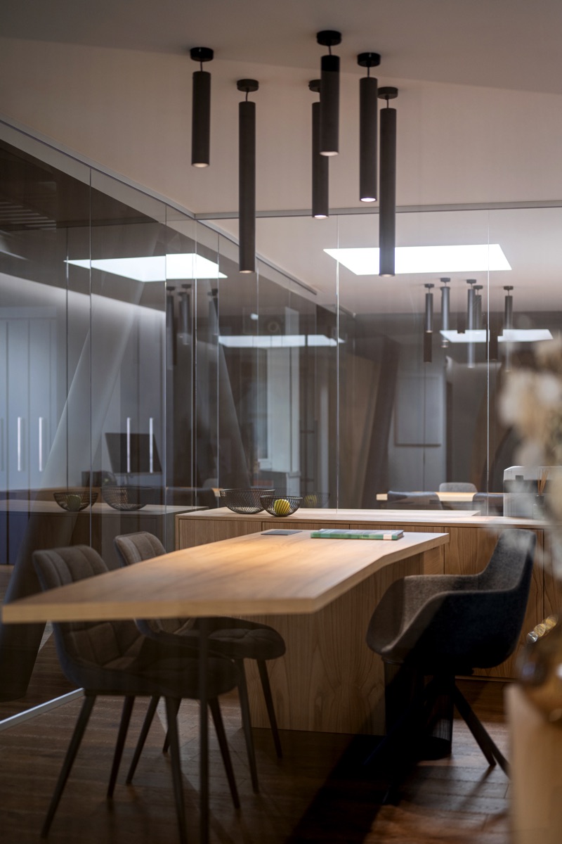 Una sala riunioni con pareti di vetro, un tavolo in legno chiaro e sedie scure. Dal soffitto pendono lampade cilindriche nere. La trasparenza delle pareti di vetro permette di vedere altre parti dell'ufficio sullo sfondo.