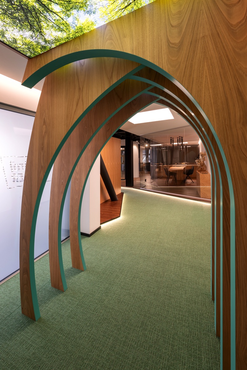 Un corridoio decorato con strutture ad arco in legno. Il soffitto ha un’immagine di foglie verdi, dando l’impressione di essere all'aperto sotto gli alberi. Il pavimento ha un tappeto verde che si estende lungo il corridoio, creando un'atmosfera naturale.