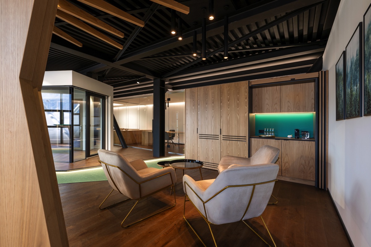 Un'area lounge moderna con poltrone beige e strutture in legno. Sullo sfondo, un mobile con illuminazione verde integrata. Il pavimento è in legno scuro, creando un contrasto elegante con l'arredamento chiaro.