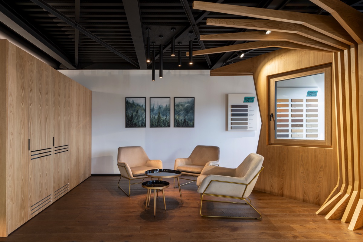 Un'area lounge con poltrone beige e strutture in legno. Sullo sfondo, tre quadri raffiguranti una foresta e un campionario di colori su una parete bianca. Il pavimento è in legno scuro, contrastando con l'arredamento chiaro.
