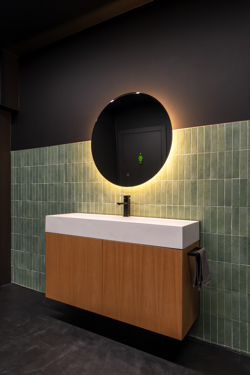  Un bagno moderno con un lavabo rettangolare bianco e mobile in legno chiaro. Lo specchio rotondo sopra il lavabo è illuminato con una luce circolare. Le pareti sono rivestite con piastrelle verdi nella parte inferiore e verniciate di nero nella parte superiore.