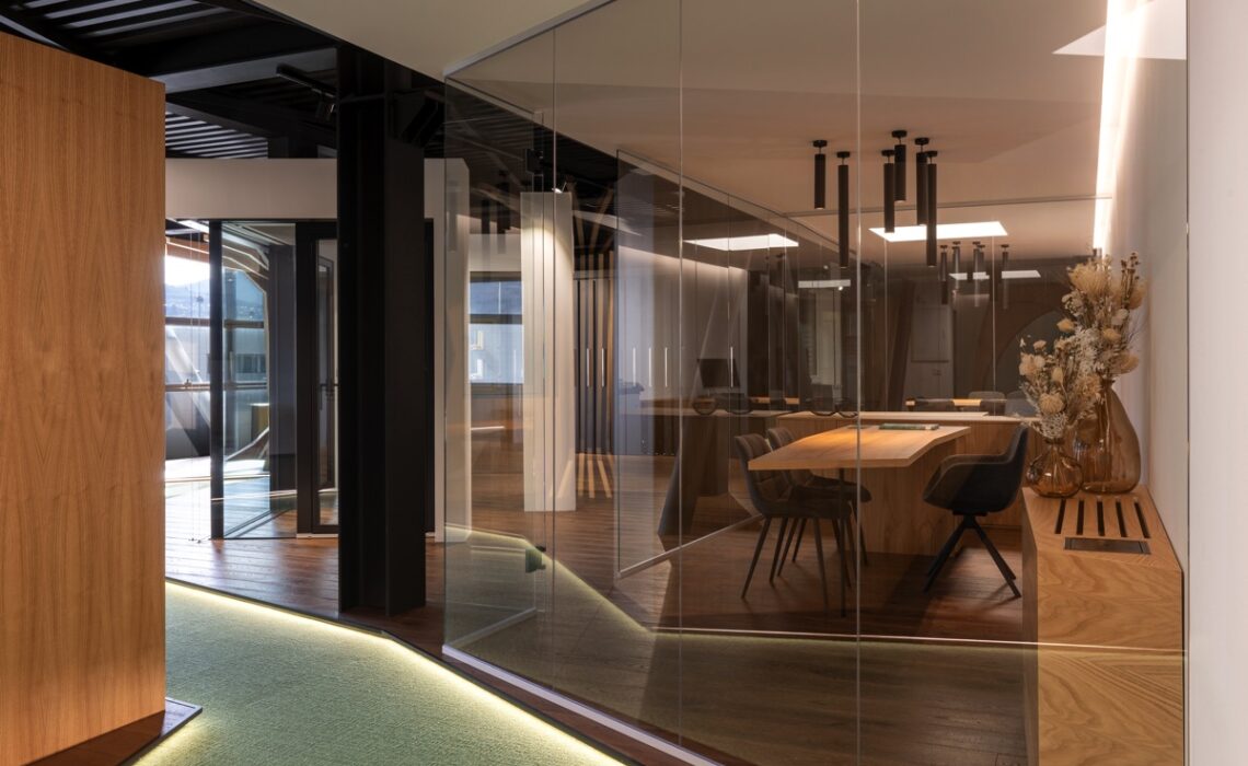 Una moderna sala riunioni con pareti di vetro, pavimento in legno scuro e sedie nere contemporanee intorno a un tavolo di legno. L'illuminazione è calda e soffusa, creando un'atmosfera accogliente. Sul lato destro, c'è una panca di legno con un vaso contenente fiori secchi. Il soffitto è dotato di moderni faretti neri. L'ambiente è professionale ma invitante, con un design elegante e minimalista.