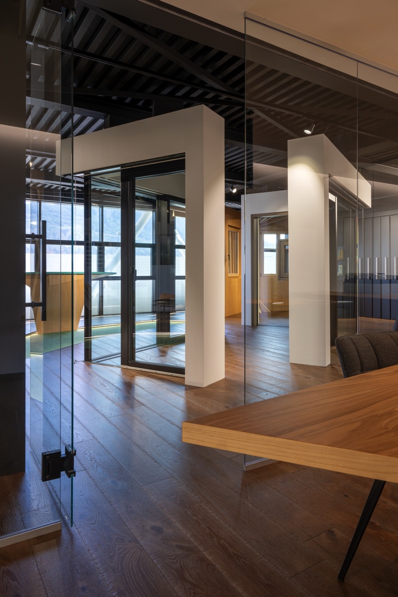 Un ufficio con pareti di vetro e strutture moderne. Il pavimento è di legno scuro e ci sono elementi in legno chiaro che contrastano piacevolmente. La luce naturale entra dalle finestre e le pareti di vetro creano una sensazione di spazio aperto e luminoso.