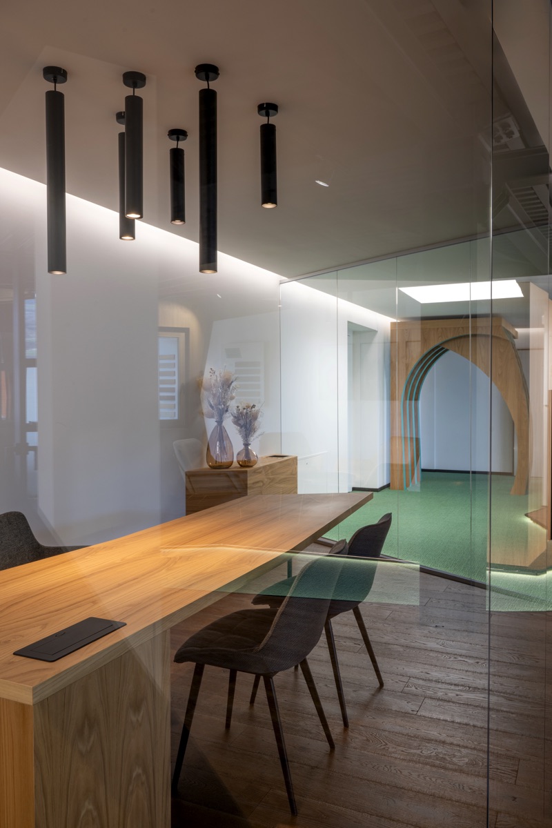 Una sala riunioni con pareti di vetro, tavolo in legno chiaro e sedie nere. Il soffitto presenta lampade pendenti cilindriche nere. Sullo sfondo, un corridoio con un arco in legno e un tappeto verde.