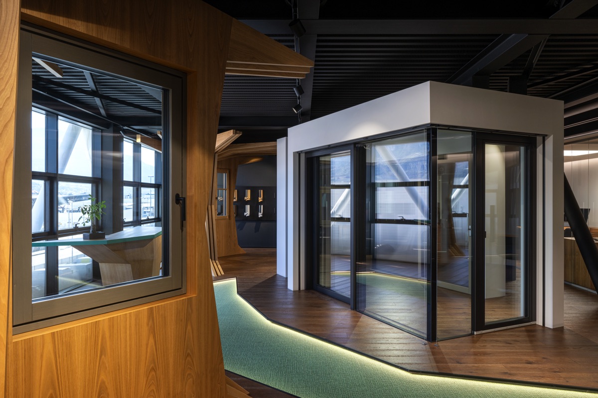 Un interno moderno con grandi finestre e strutture in legno. Il pavimento è di legno scuro con un percorso verde illuminato. Le pareti di vetro creano un ambiente aperto e luminoso, accentuato dalla luce naturale che entra dalle finestre.