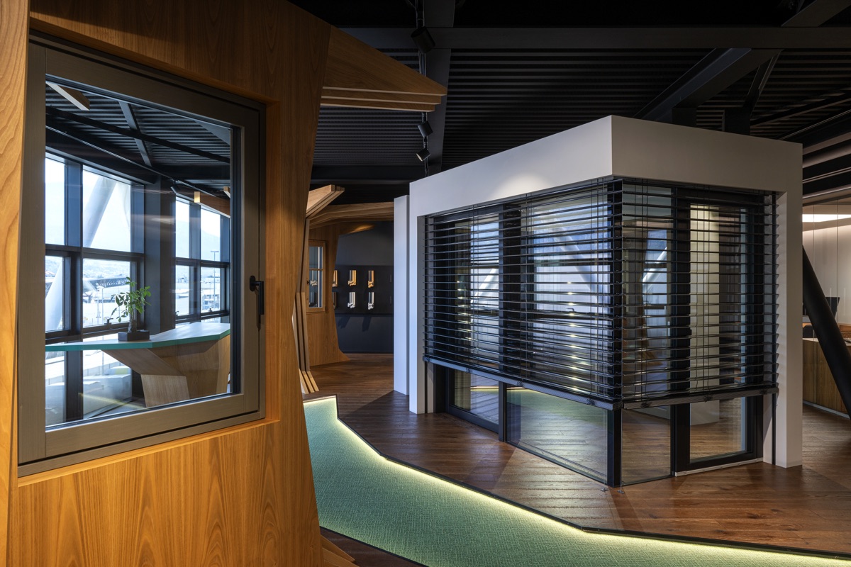 Interno moderno con grandi finestre e strutture in legno. Il pavimento è di legno scuro con un percorso verde illuminato. Una sezione dell'ufficio ha una schermatura in legno e vetro, creando una divisione elegante e funzionale.