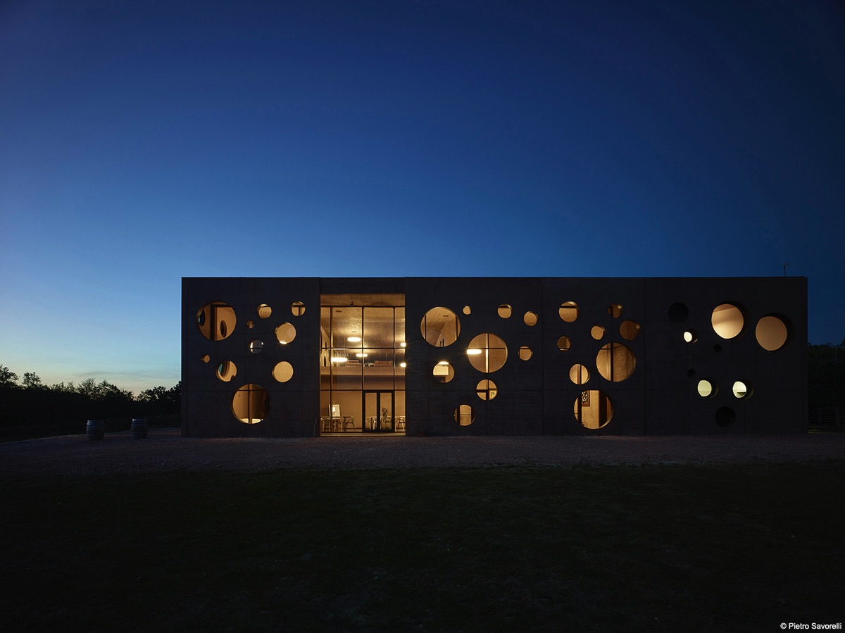 Cantina del Podere la Chiesa, struttura moderna con facciata caratterizzata da aperture circolari di varie dimensioni, illuminata di sera.